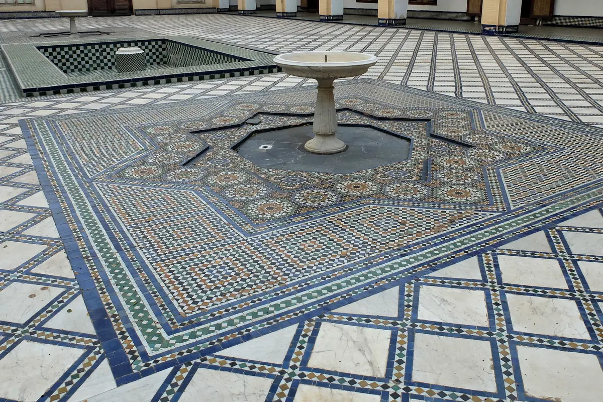 A courtyard floor of zellij mosaic in green, blue and ochre around a stone fountain, with a tiled basin beyond it