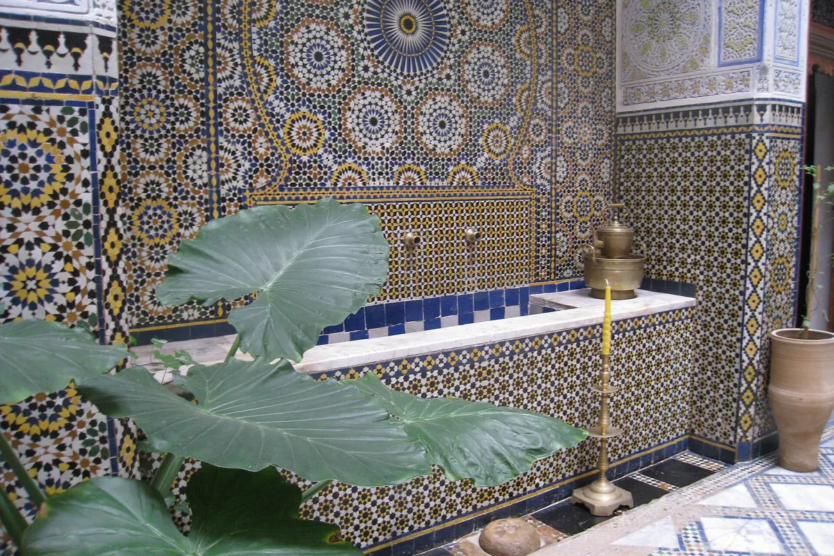 En fontaneniche dækket af geometrisk zellij-mosaik i et riad-hus i Fes, med et stort grønt blad i forgrunden.