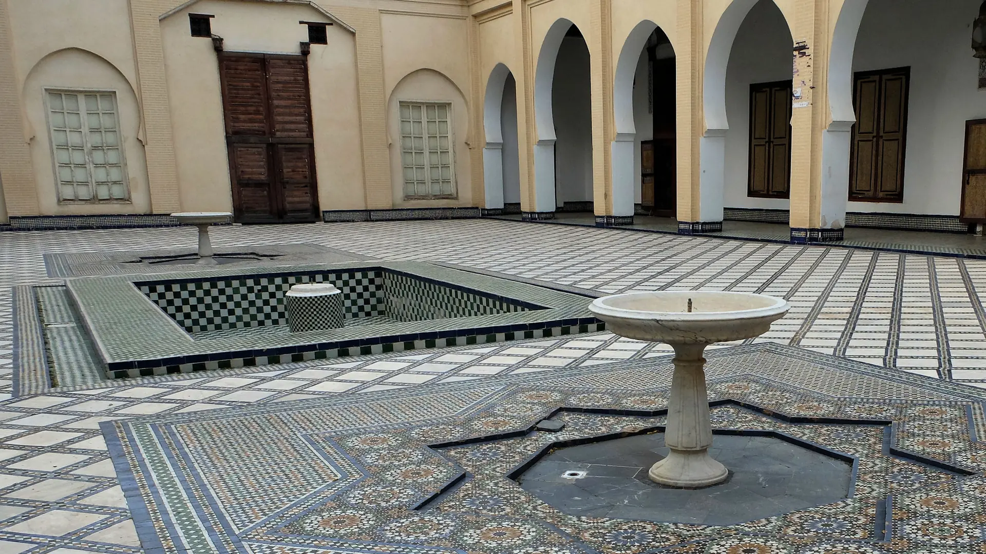 En gårdsplads i et historisk palads i Fes, belagt med zellij-fliser og med en stenfountain i midten, under en hvid arkade af hesteskoformede buer.